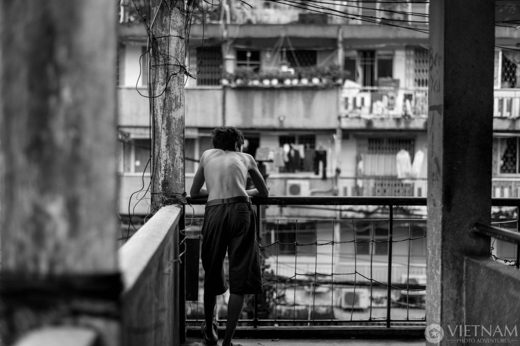 A black and white candid photography of a shirtless Vietnamese man, seen for the back, leaning on a balcony of an old Saigon building.