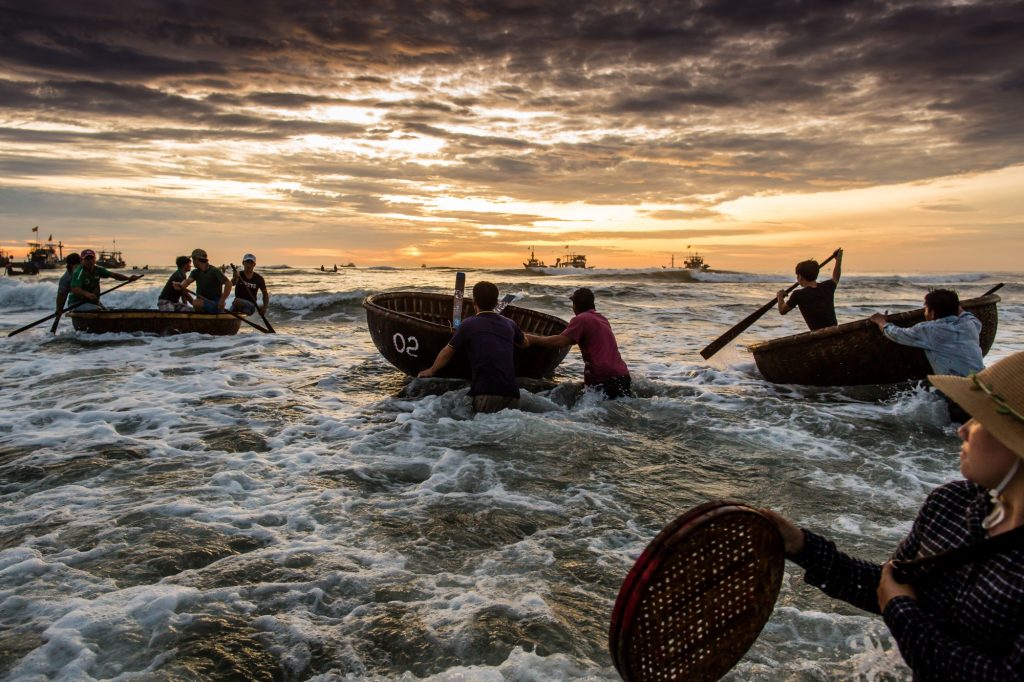 Candid photography of a woman with a handwoven basket on the beach, facing the open sea under a dramatic dawn sky, watching a group of fishermen in three round traditional boats paddling towards the horizon, central vietnam.