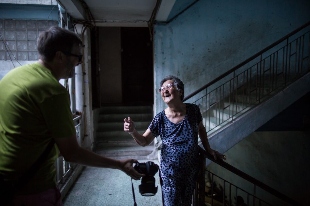 A man is showing his camera to an old Vietnamese woman in an old Saigon building interior, both laughing.