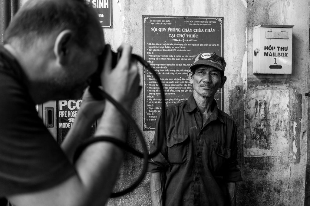 A black and white picture of a man capturing a close up portrait of a stern Vietnamese working man wearing a hat, against an urban wall full of Vietnamese writting.