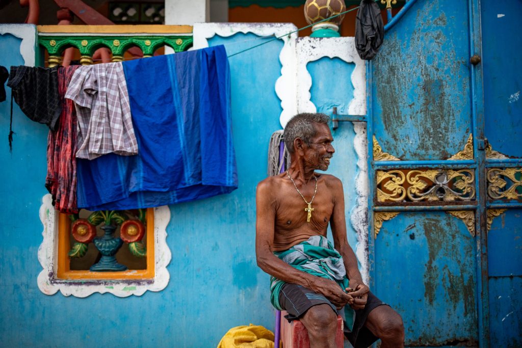 Candid street photography portrait of man in Jaffna alley, sitting on a chair shirtless with golden cross around his neck, with a large contrasting blue wall backdrop.