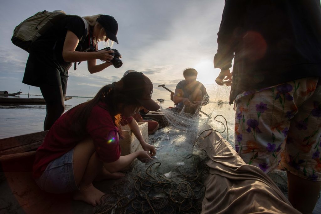 A female photographer shooting downwards from inside a small boat, capturing a group of three fishermen working on mending their nets.