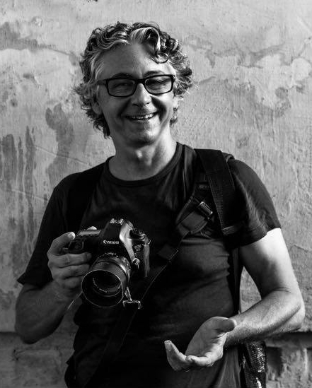 A black and white portrait of Arnaud Foucard, camera in hand, looking to the side and smiling.